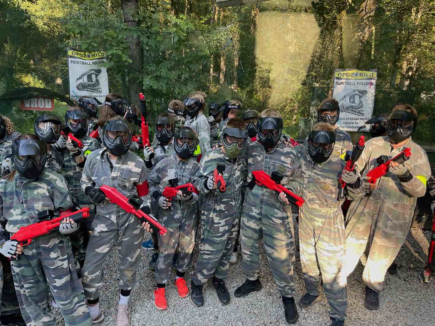ekipni Paintball za otroke Ljubljana . Prikazana je ekipa otrok v v kamuflažni obleki z zaščitno masko na obrazu in vsi imajo v rokah pištolo za paintball. Pištole so v rdeči barvi.